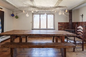 Une grande table en bois, des bancs, une cheminée en briques et une fenêtre laissant entrer la lumière naturelle.
