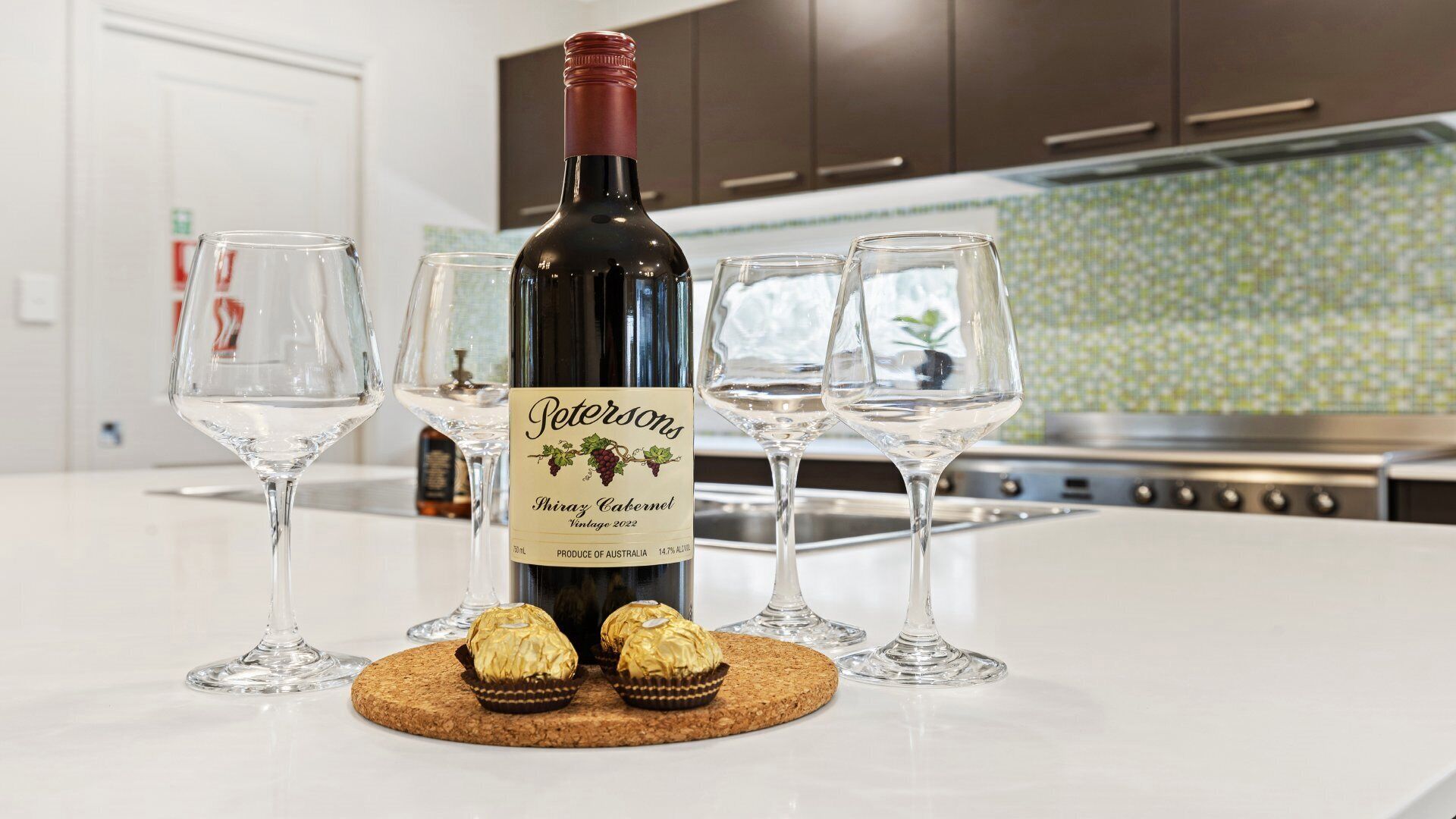 A bottle of Petronsky Shiraz Cabernet on a kitchen counter with two glasses and two chocolates on a coaster.