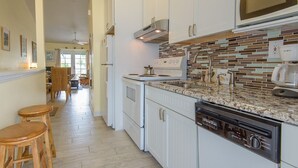A kitchen with white cabinets, a microwave, a stove, and a dishwasher. There are wooden stools and a dining area in the background. - Tropical Breezes at The Truman Annex in Key West (Key West)