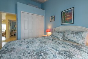 A bedroom with a bed, a nightstand, a lamp, a door, and a painting on the wall. - Tropical Breezes at The Truman Annex in Key West (Key West)