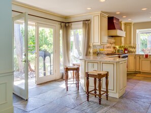 A kitchen with a large island, wooden stools, and a view of greenery outside.
