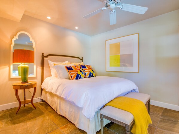 A bedroom with a bed, bedside table, lamp, bench, ceiling fan, and artwork on the wall.