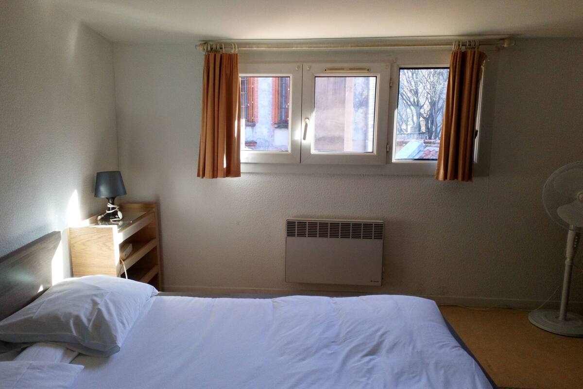 Une chambre à coucher avec un lit, une table de chevet avec une lampe, une fenêtre avec des rideaux, un radiateur et un ventilateur sur pied.