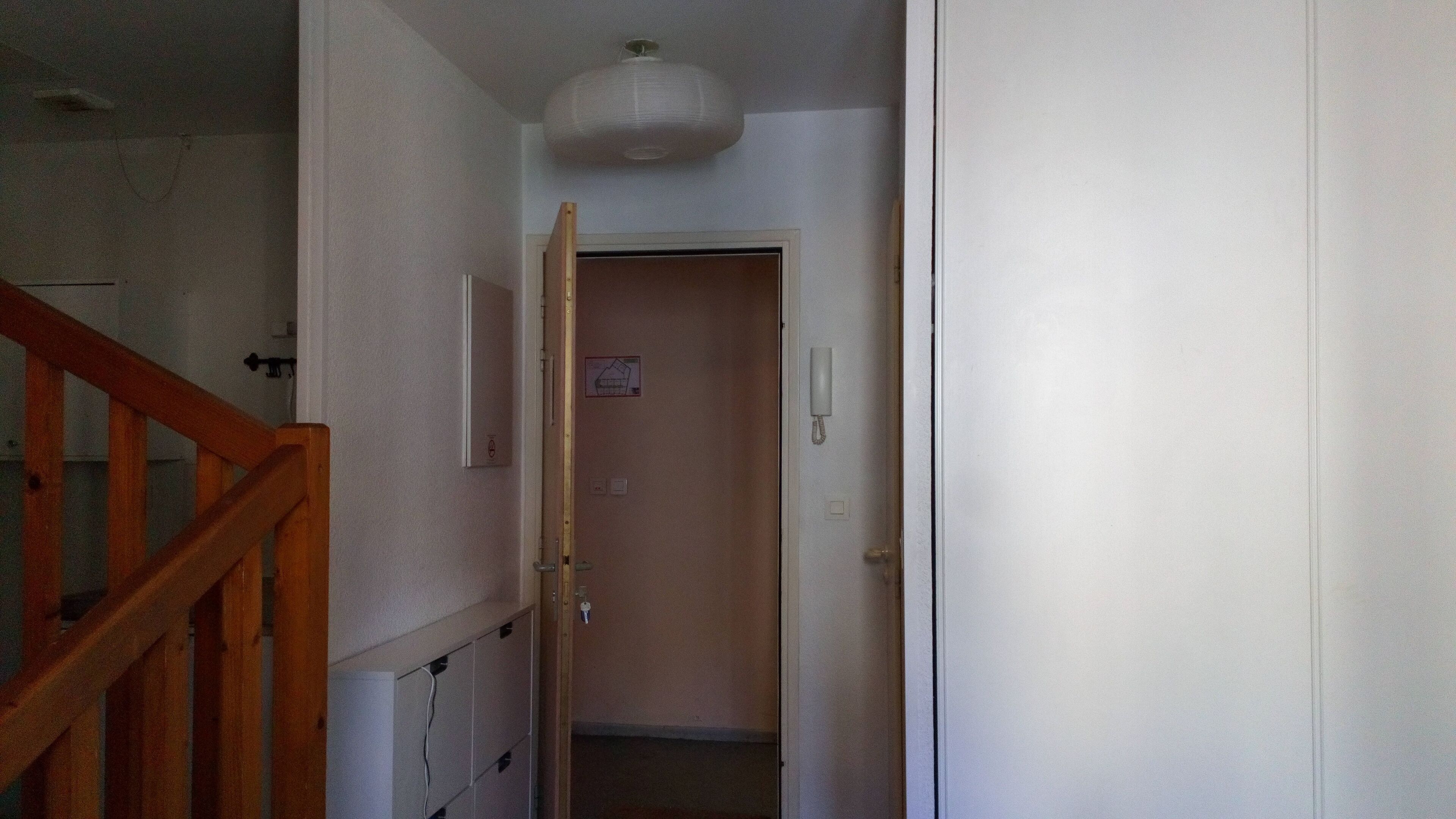 Un pasillo con una escalera de madera a la izquierda, una puerta blanca con un teléfono a la derecha y una lámpara arriba.