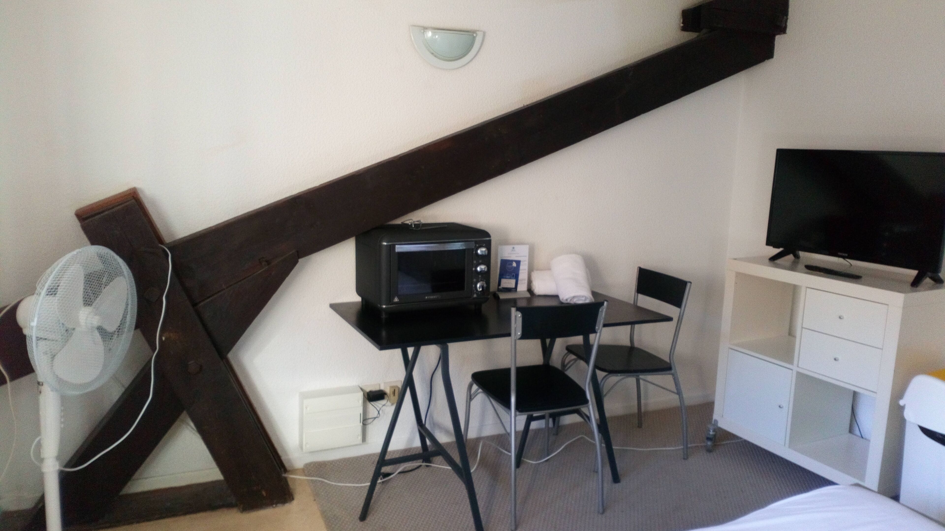 Une chambre d’hôtel compacte équipée d’une kitchenette, d’une télévision, d’un ventilateur et d’un petit bureau avec une chaise.