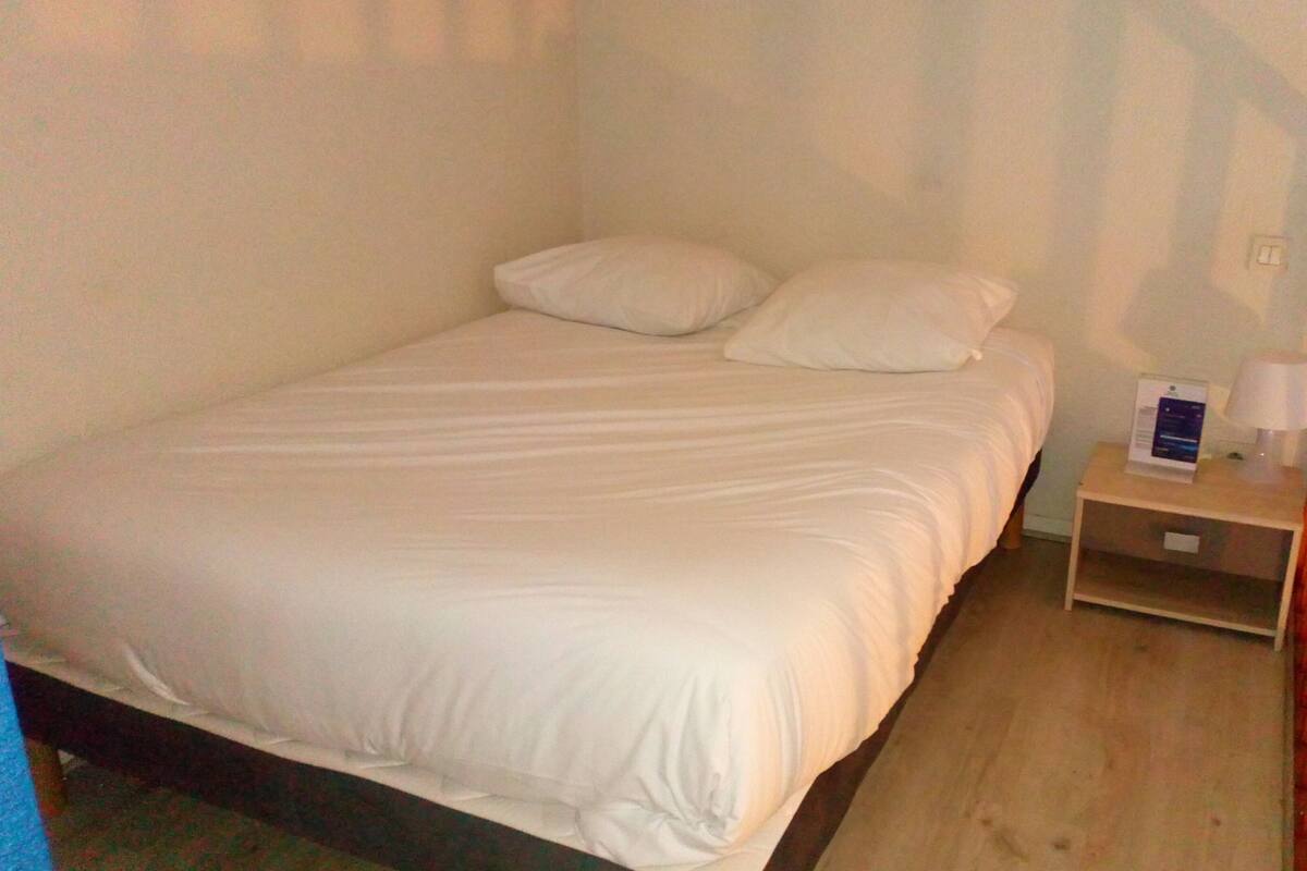 Un lit simple avec une literie blanche, dans une chambre dotée d’une tête de lit en bois et d’une table de chevet sur laquelle se trouvent une lampe et une brochure.