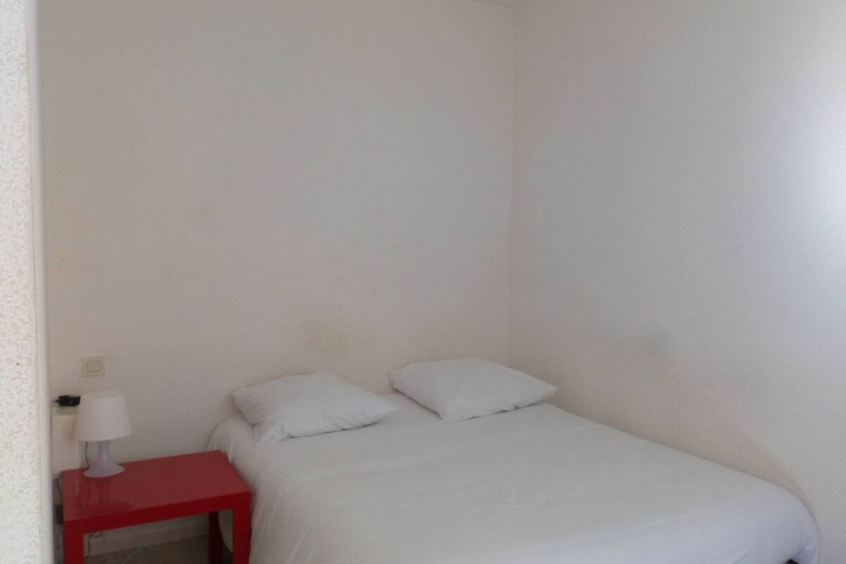 Une petite chambre blanche avec un lit simple, une table de chevet rouge et une lampe blanche.