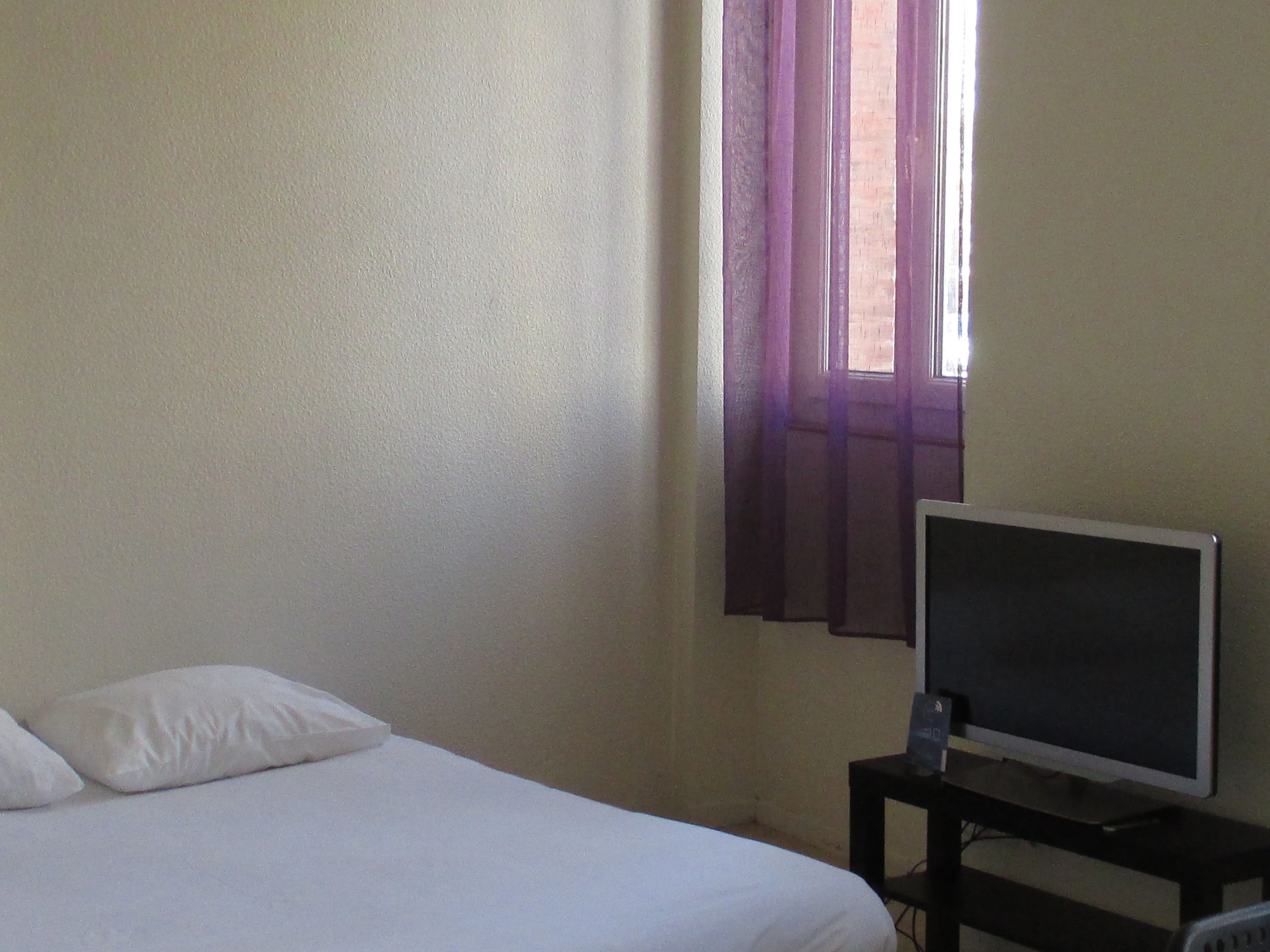 A simple room with a bed, a television on a stand, and a window with purple curtains.