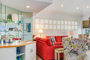 A modern living room with a red sofa, a glass coffee table, and a floral armchair. There is a kitchen area with a wooden countertop and a glass cabinet.