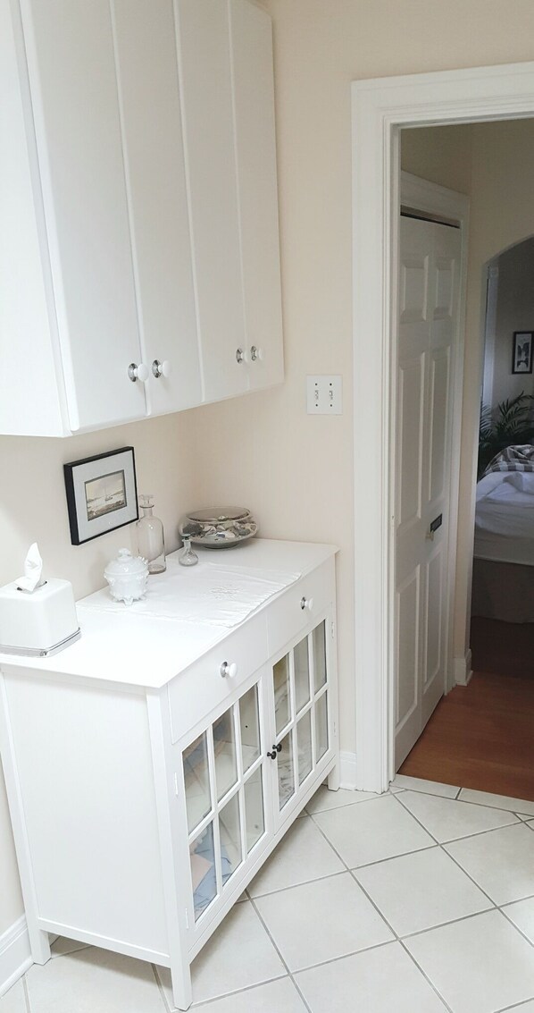 A white bathroom with a vanity, a framed picture on the wall, and a glimpse of a bedroom with a bed.
