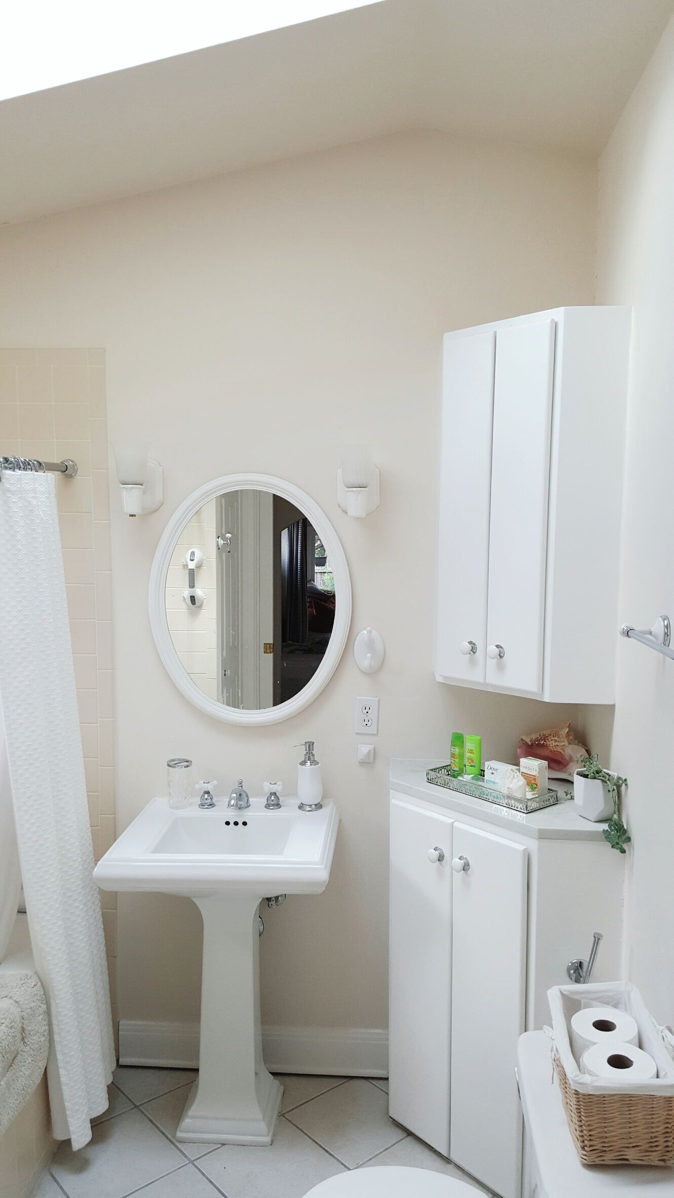 Een badkamer met een wastafel op voetstuk, een ronde spiegel, een wit kastje en een rieten mand met toiletpapier.