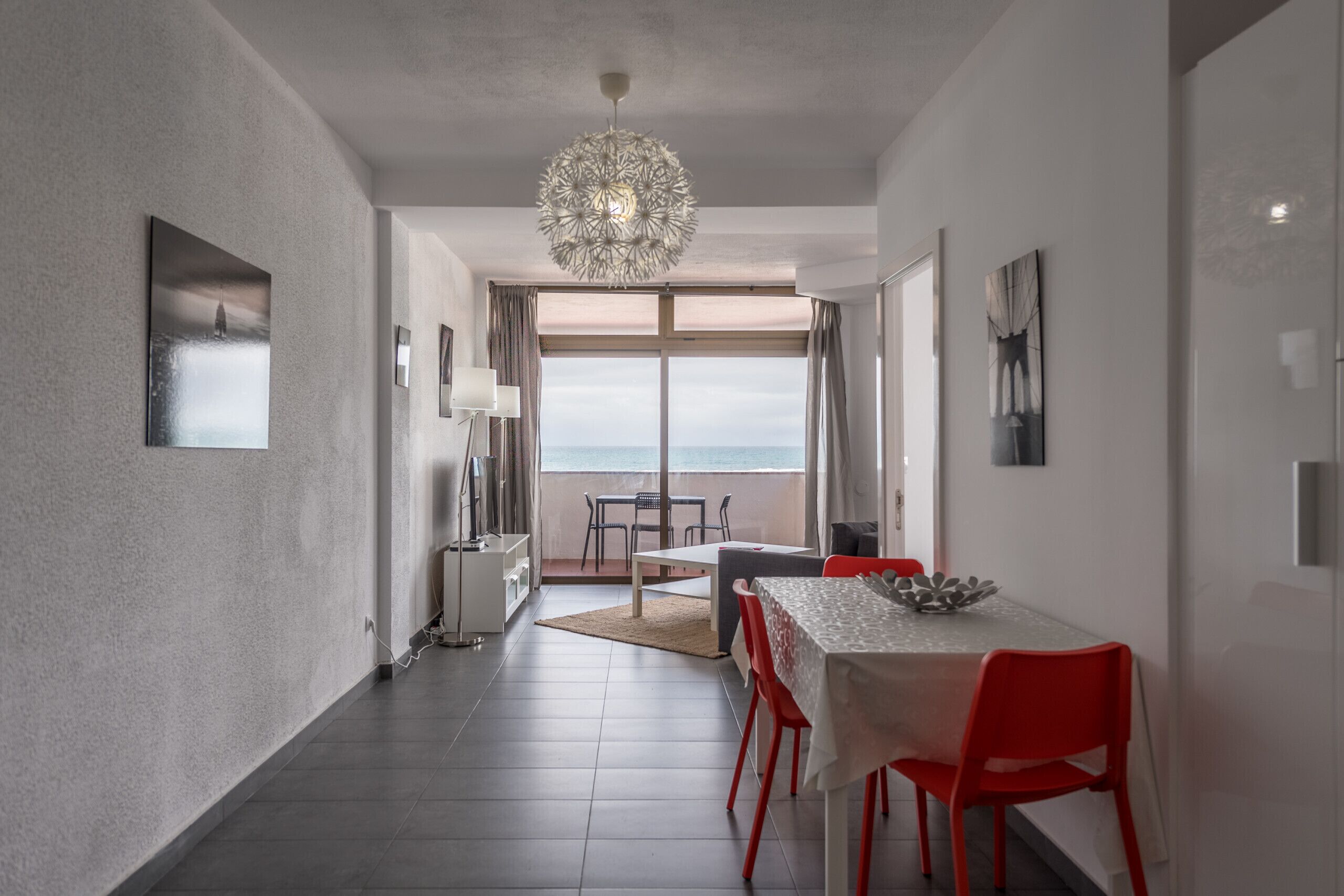 Un comedor moderno con una mesa, sillas, una araña y vistas al mar a través de amplios ventanales.