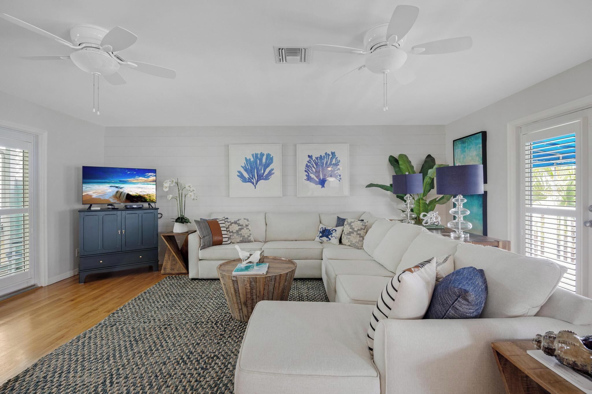 A modern living room with a sofa, coffee table, and a TV. There are ceiling fans, artwork on the walls, and a window with blinds.