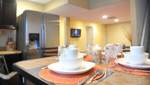 Un área de comedor con una mesa lista para dos personas, con cuencos y tazas blancos, una vela y un televisor al fondo.