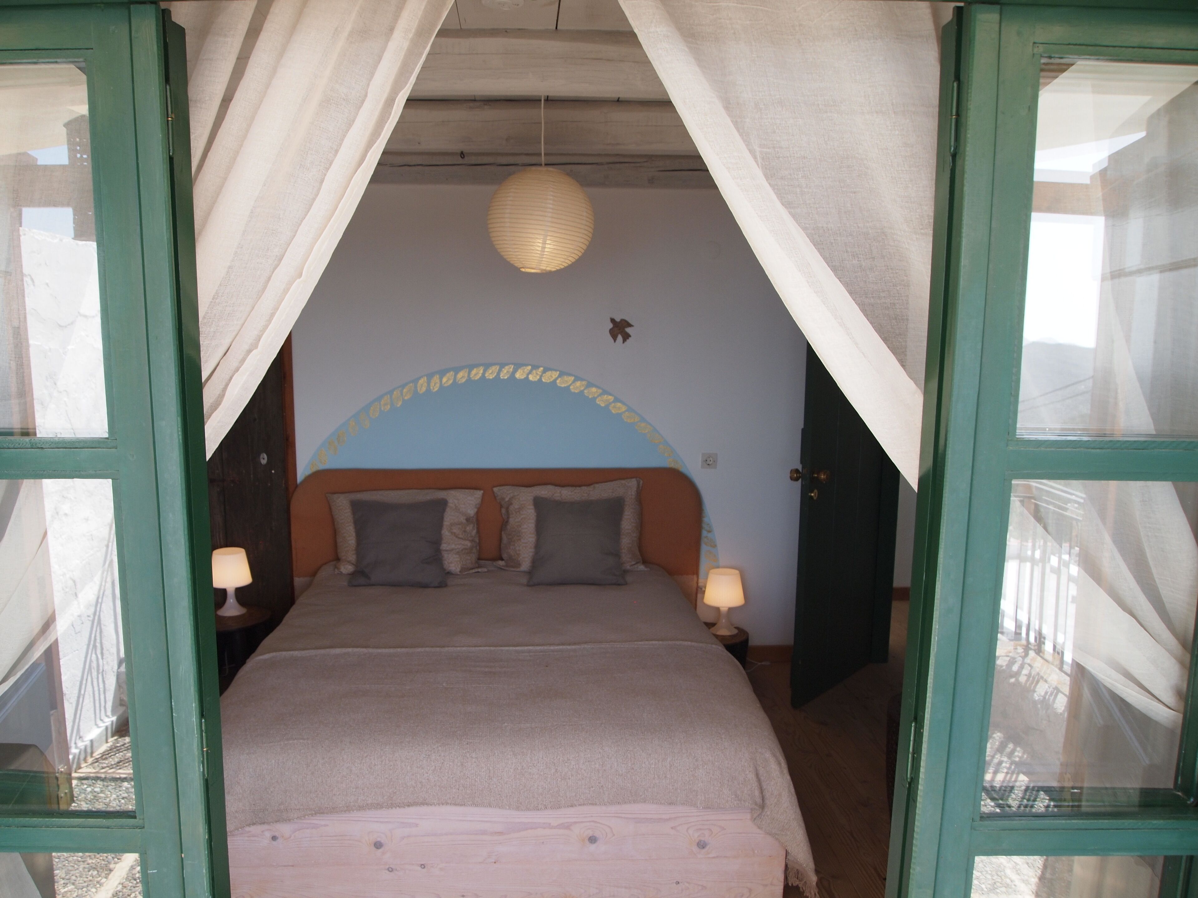 A bedroom with a bed, two bedside lamps, a hanging light fixture, and a door leading to another room.