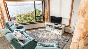 A living room with a large window offering a view of the water, a flat-screen TV on a white stand, and a grey shag rug. - Larus Waterfront Cottage (White Beach)