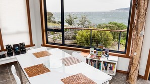 A modern dining area with a large window overlooking the sea, a white table with woven placemats, and a bookshelf filled with books. - Larus Waterfront Cottage (White Beach)