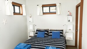 A neatly arranged bedroom with a striped bedspread, two pillows, a window with curtains, and a small shelf with a decorative item. - Larus Waterfront Cottage (White Beach)