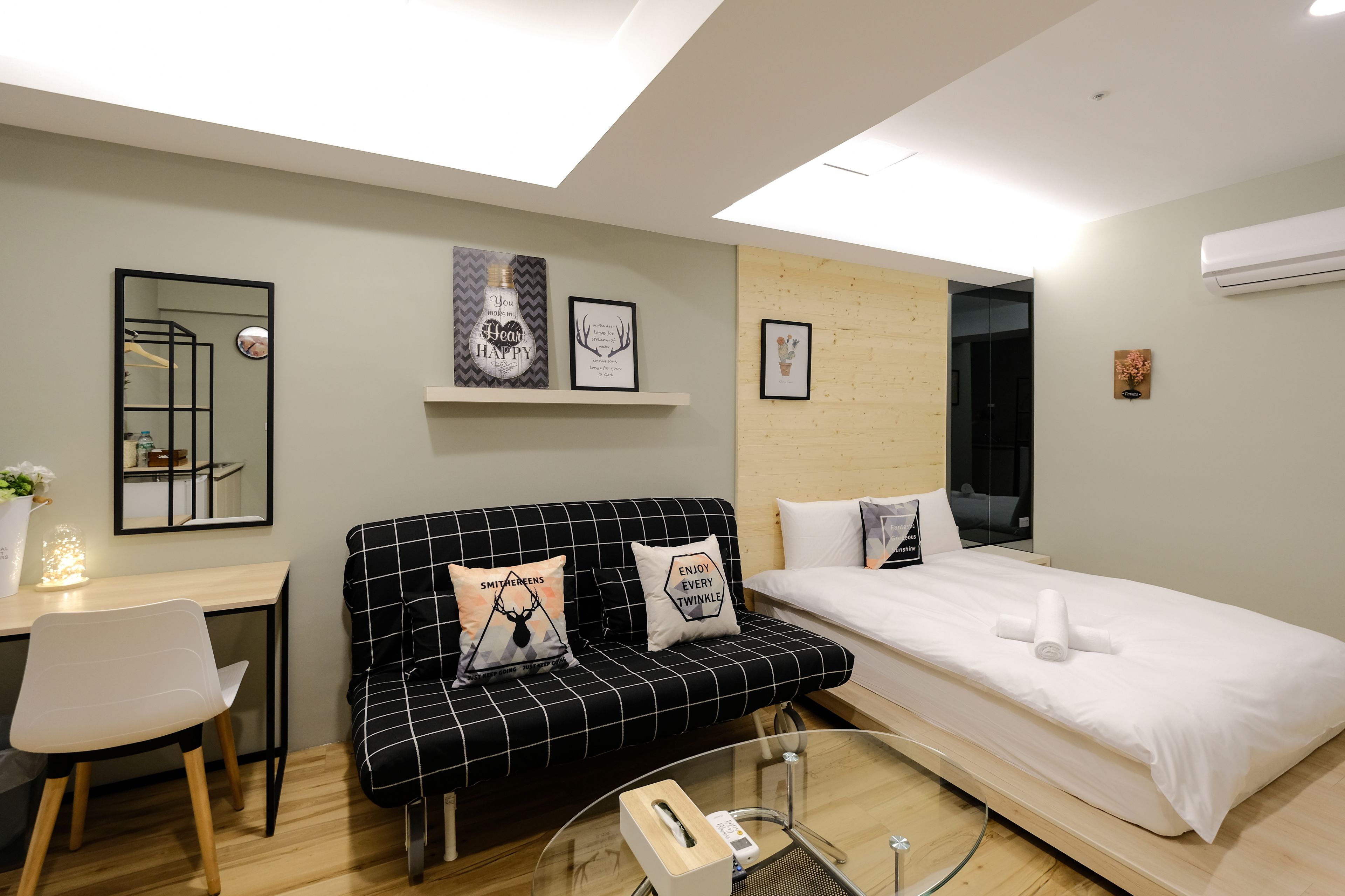 A modern hotel room with a black and white checkered sofa, a single bed, a small desk with a chair, and a mirror.