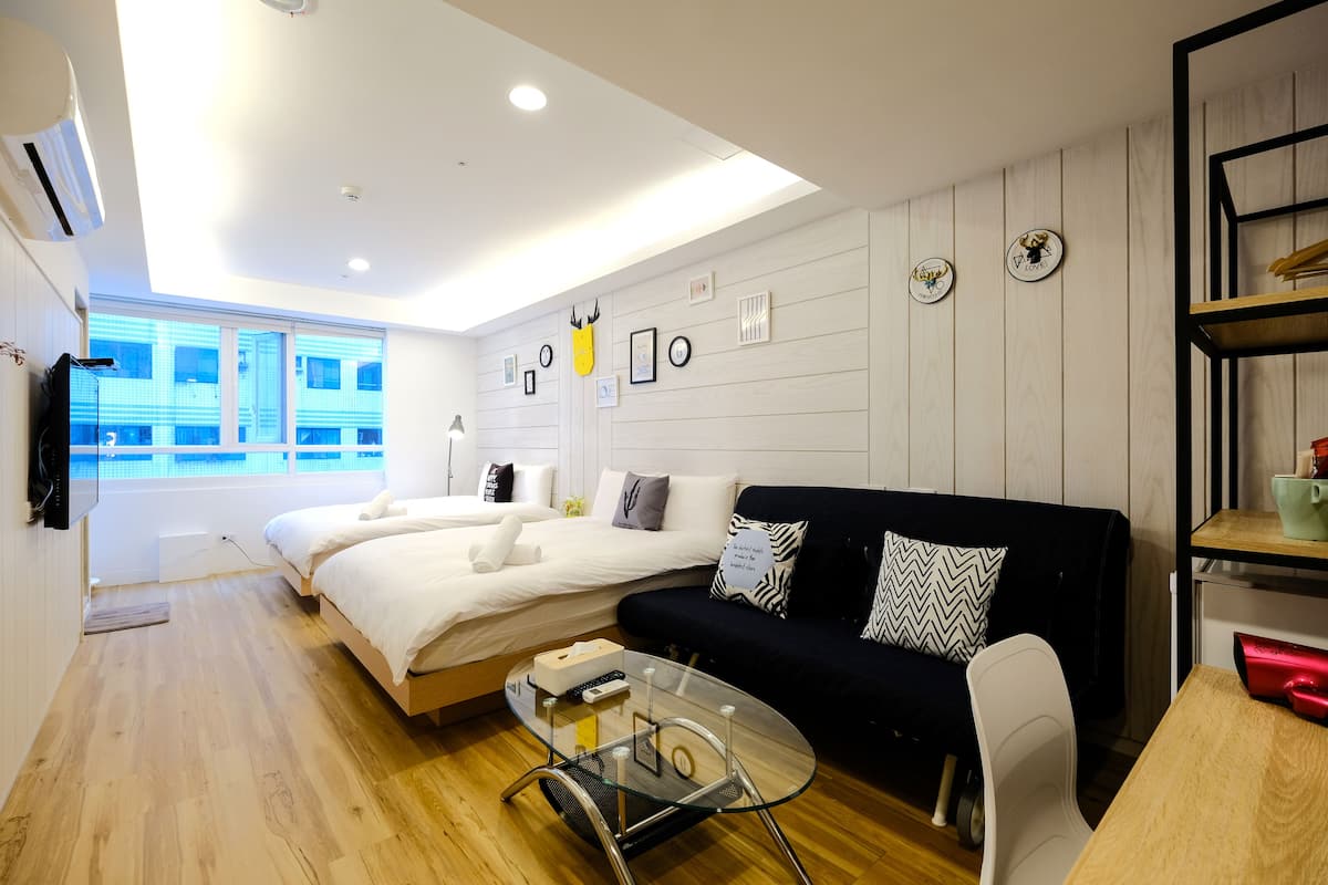 Quarto de hotel moderno com uma cama grande, um sofá preto, uma mesa de centro de vidro e piso de madeira.