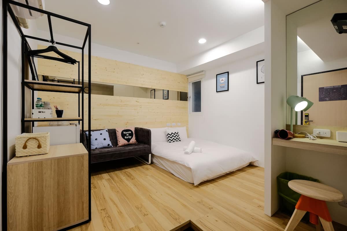 Quarto de hotel moderno com piso de madeira, cama, sofá, escrivaninha, prateleira suspensa e um pequeno banquinho.