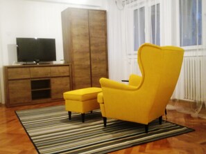 Un salon avec un fauteuil jaune, un petit pouf, un meuble en bois et un téléviseur posé sur un meuble.
