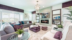 A modern living room with a grey sofa, a white armchair, and a glass coffee table. There is a fireplace, a chandelier, and artwork on the walls. - Spacious Villa with Home Theater at Encore (Kissimmee)