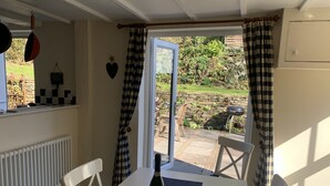 Un comedor con una mesa dispuesta para dos personas, una botella de vino y una copa. Un ventanal con cortinas a cuadros da a un jardín con una parrilla.