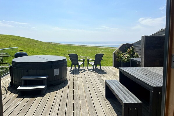 A wooden deck with a hot tub, two chairs, and a table overlooking a grassy landscape.