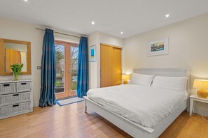 A bedroom with a large bed, a wooden wardrobe, a mirror, a door to a balcony, and a small bedside table with a lamp.