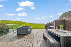 A wooden deck with a hot tub, outdoor seating, and a scenic view of a green hillside and ocean.