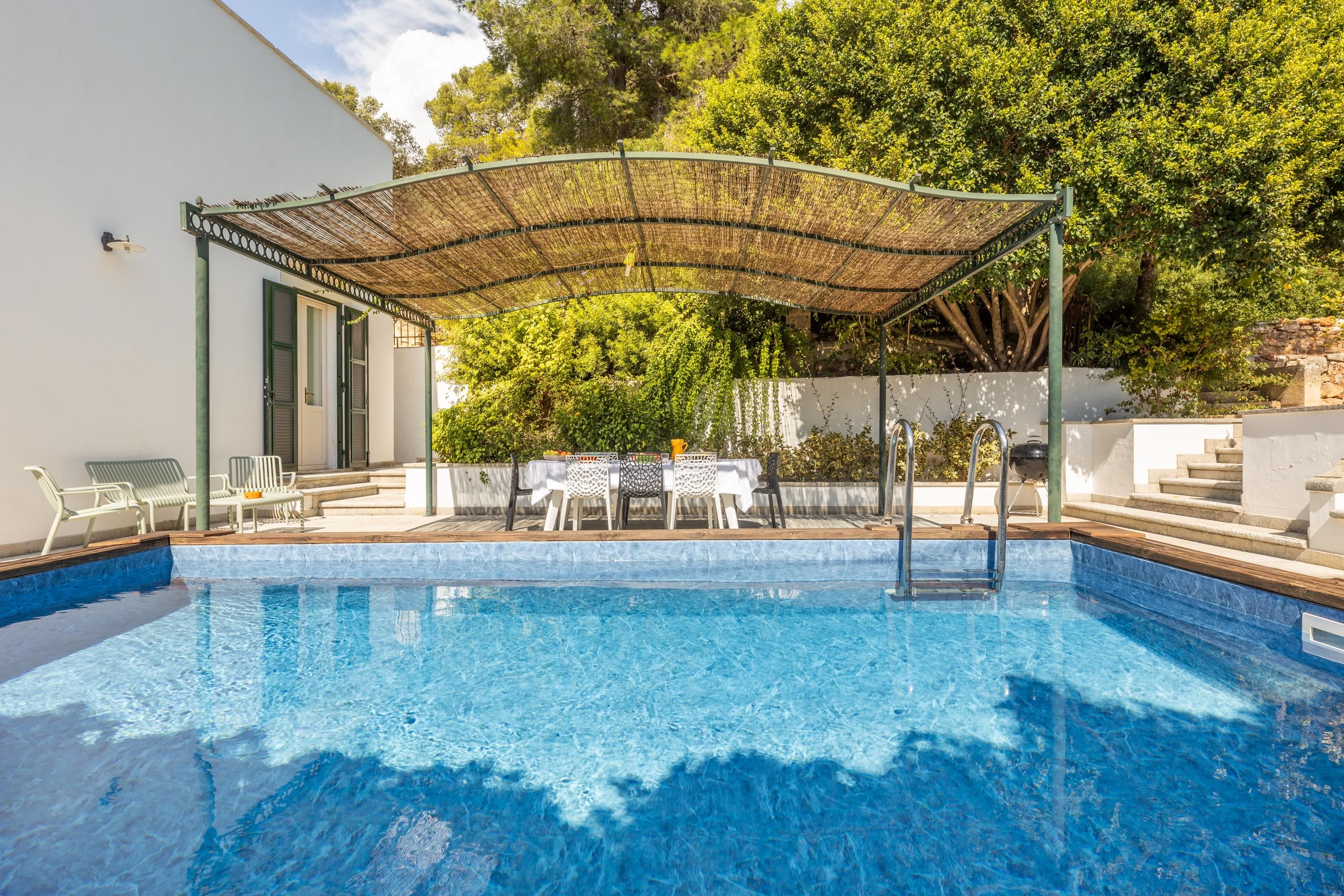 Una piscina con un patio coperto, posti a sedere all'aperto e vista su alberi e un edificio.