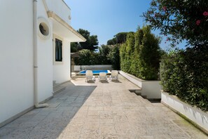 A pool area with a table and chairs, surrounded by greenery and a white building.