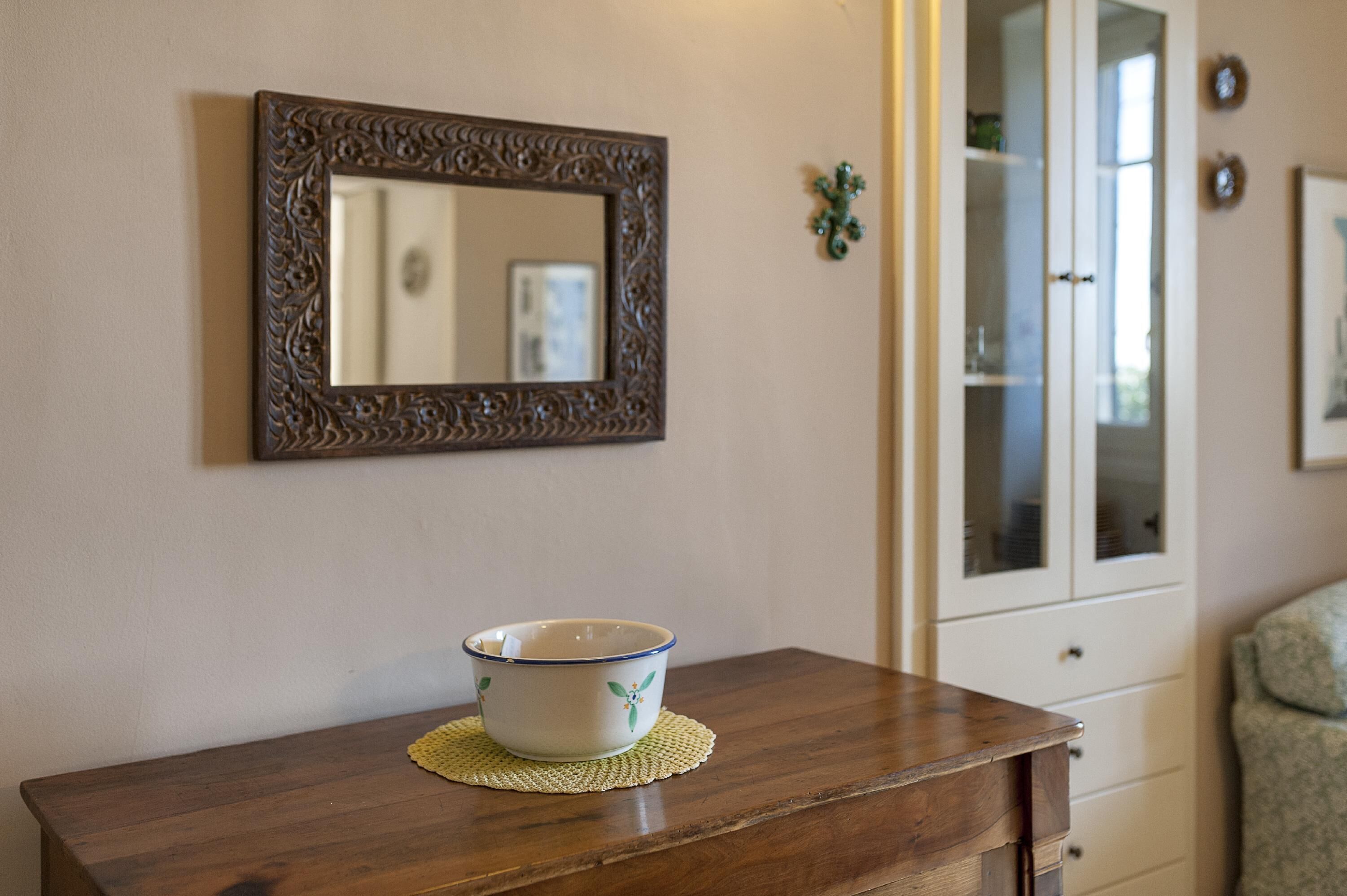 A wooden table with a bowl on a placemat, a mirror with an ornate frame, and a cabinet with glass doors in the background.