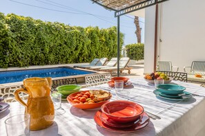 A poolside dining area with a table set for two, surrounded by lounge chairs, and a view of a swimming pool.