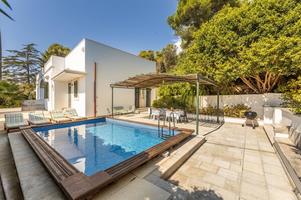 A modern outdoor pool area with a pergola, lounge chairs, and a barbecue grill.