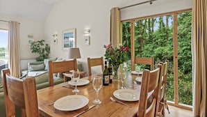 A dining area with a wooden table set for four, a vase with flowers, and a view of greenery through large windows. - Beautiful holiday home with hot tub, just a walk away from Talland beach... (Looe)
