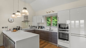 A modern kitchen with a central island, stainless steel appliances, and a large window with a view of greenery. - Beautiful holiday home with hot tub, just a walk away from Talland beach... (Looe)