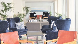 Un área de estar moderna con sillones azul oscuro, un sofá central gris y una pequeña mesa. Hay plantas en macetas y un gran ventanal que ofrece vistas al exterior.