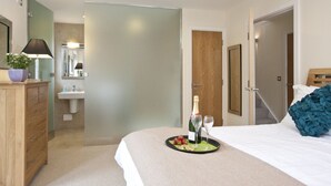 A hotel room with a bed, bedside table, a nightstand, a dresser, a mirror, a door, and a bathroom visible through a frosted glass partition. - Beautiful holiday home, just a walk away from Talland beach... (Looe)