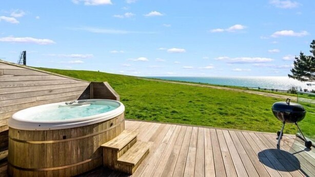 A wooden deck with a hot tub and a grill, overlooking a grassy area and the ocean. - Beautiful holiday home, just a walk away from Talland beach... (Looe)