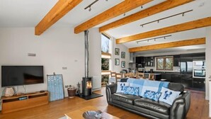 A living room with a fireplace, a sofa, a TV, and a kitchen in the background. - Beautiful holiday home, just a walk away from Talland beach... (Looe)