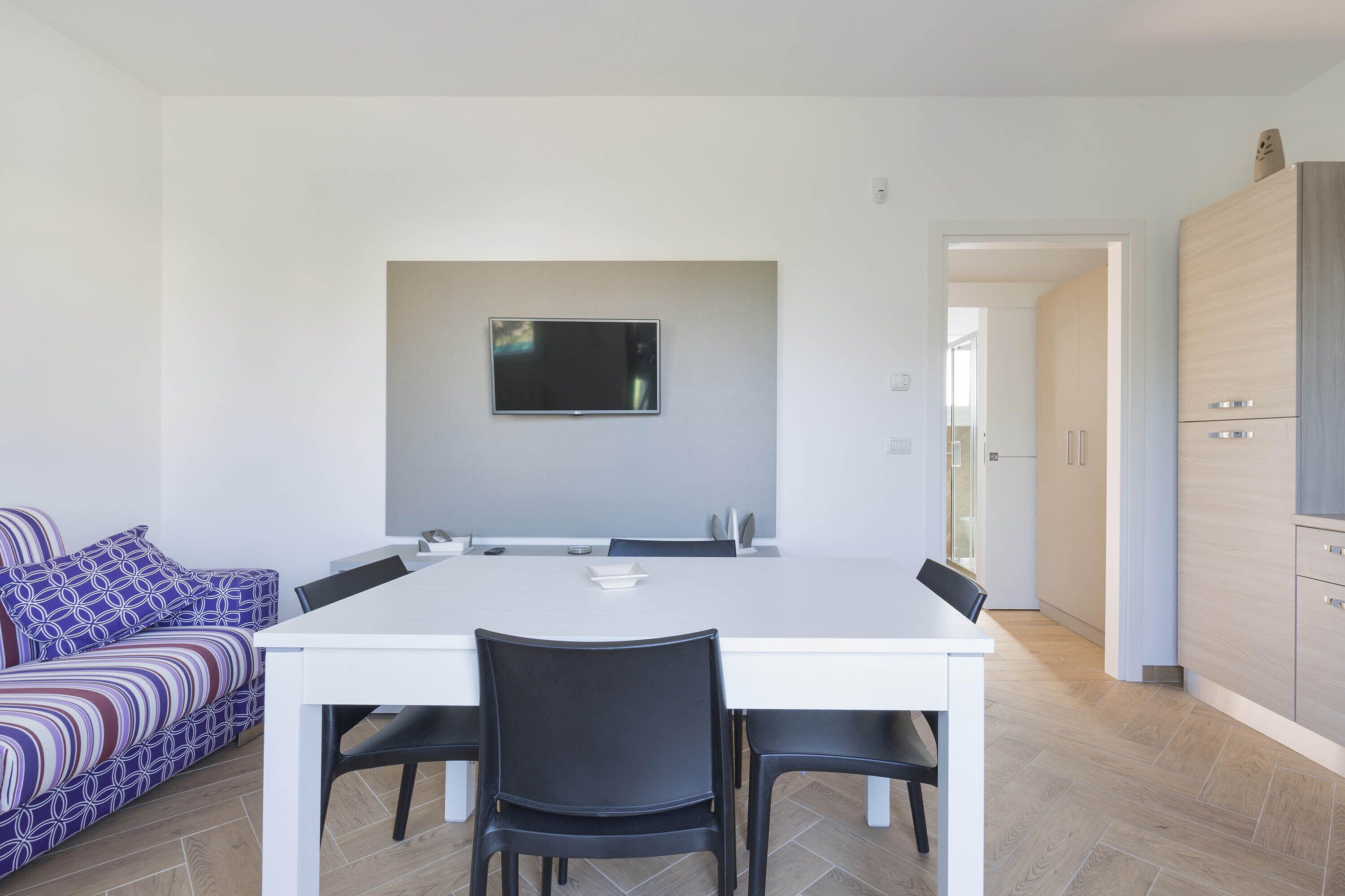 A modern kitchen with a white table and chairs, a flat-screen TV mounted on the wall, and a patterned sofa in the background.
