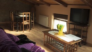 Une pièce mansardée chaleureuse avec un plafond en bois, une table à manger avec des chaises, un téléviseur posé sur un meuble et une fenêtre avec des rideaux.