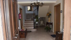 Un couloir avec une porte en bois, un extincteur, un escalier, un lustre et un vase rempli de fleurs.