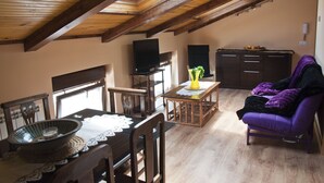 Une pièce aux hauts plafonds, avec un plafond en bois et un puits de lumière, une table à manger agrémentée d’un saladier et d’un vase, un canapé violet et un coin cuisine avec des placards et un téléviseur.