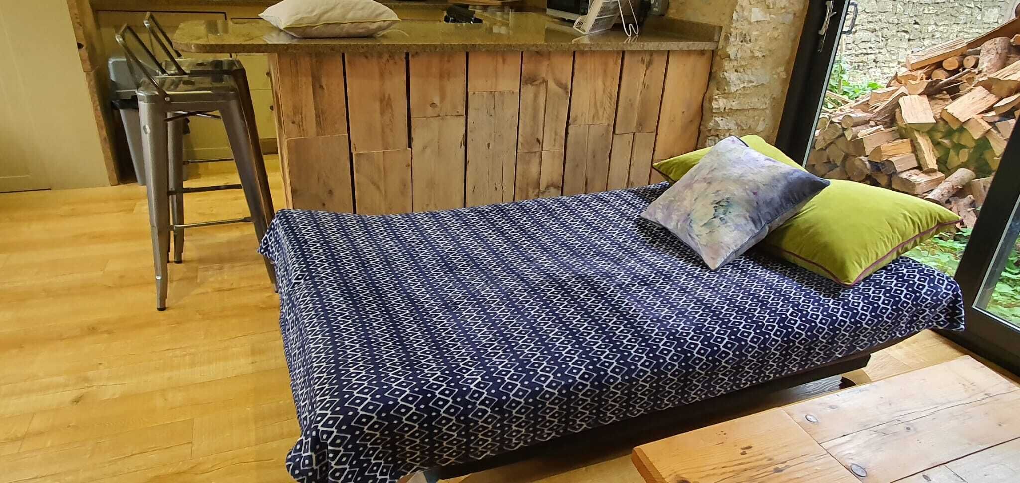 Ein Einzelbett mit einem gemusterten Bettüberwurf, einem grünen Kopfkissen und einem Holz-Kopfteil in einem Zimmer mit Holzboden und Steinwand.