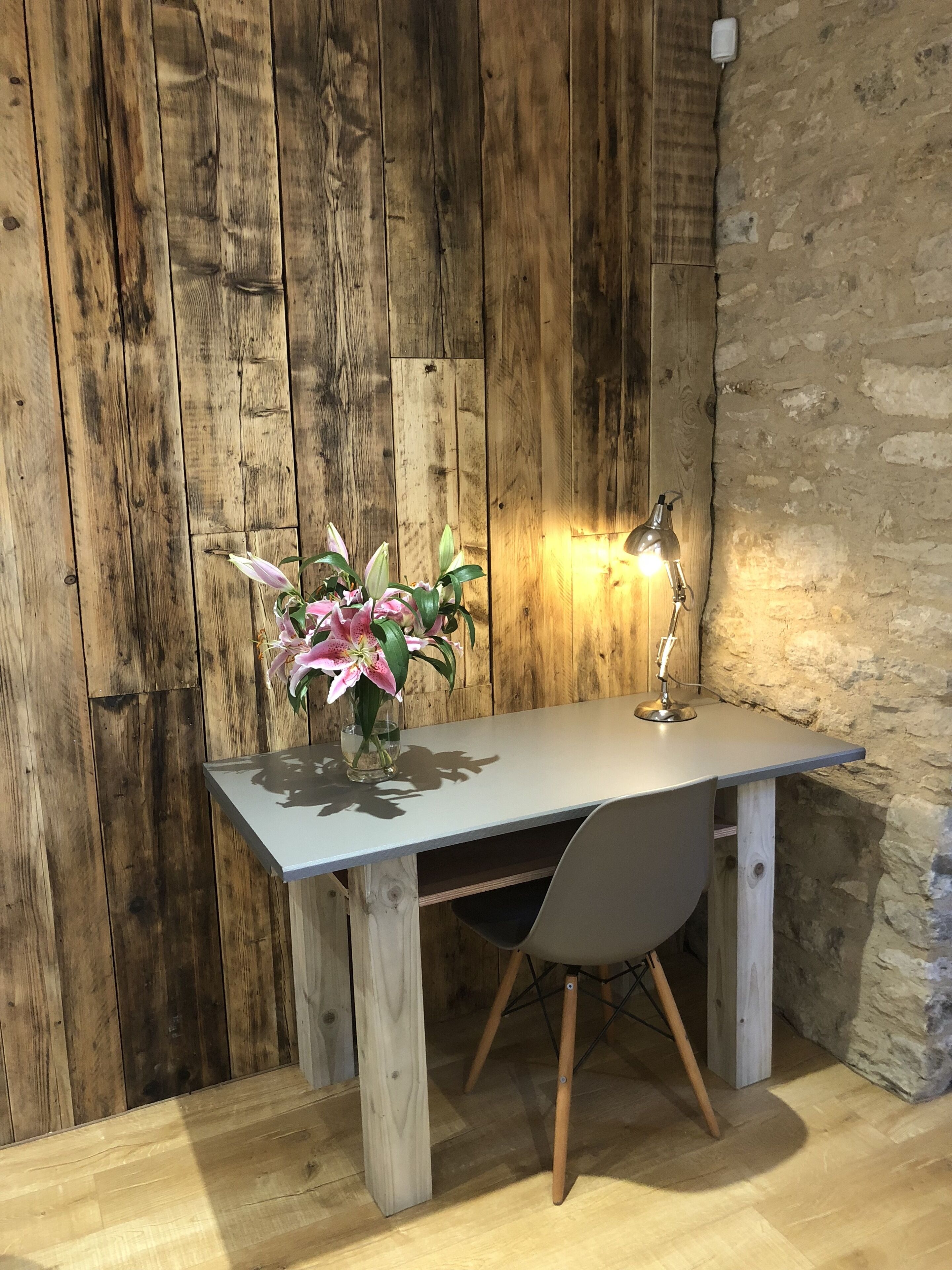 A wooden desk with a vase of flowers and a desk lamp against a rustic wall.