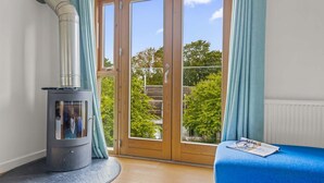 Ein Zimmer mit Kaminofen, einem blauen Sofa und Blick auf Bäume durch eine Glastür.