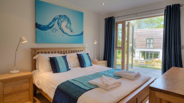 Ein Schlafzimmer mit einem großen Bett, einem Nachttisch, einer Nachttischlampe, einem Gemälde einer Welle und Blick auf ein Gebäude durch eine Glastür.